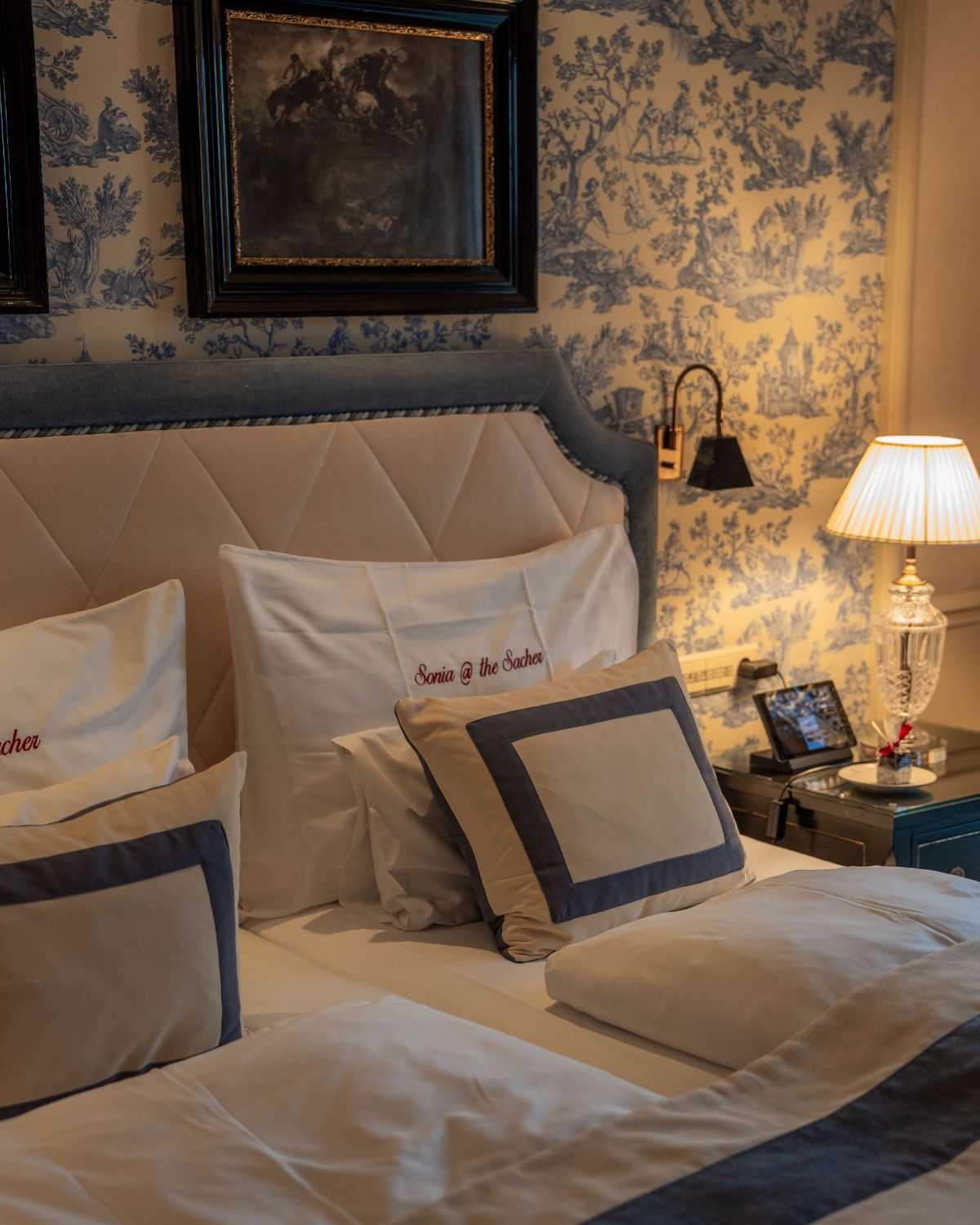 A hotel room setting with blue and white wall paper, bedding, and a crystal table lamp.