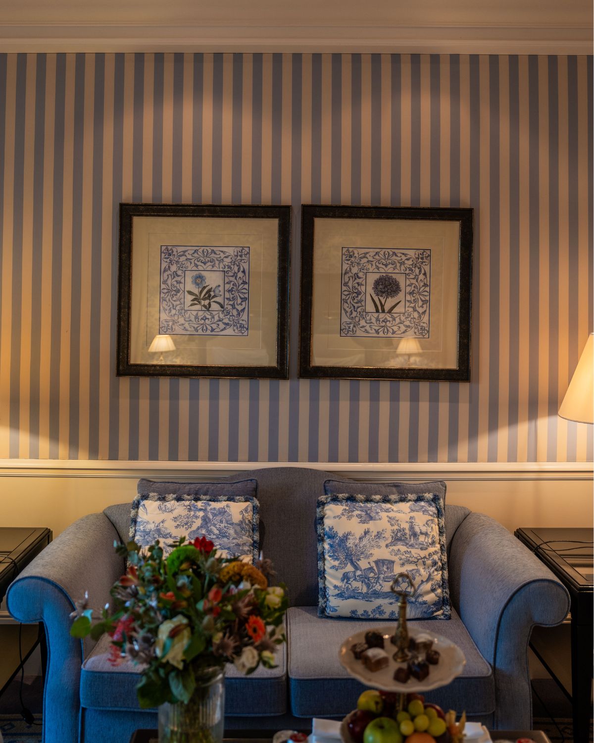 A blue and white stripe wallpaper with gold frames, a blue sofa next to two dark side tables and a center table with flowers, sweets and fruits.