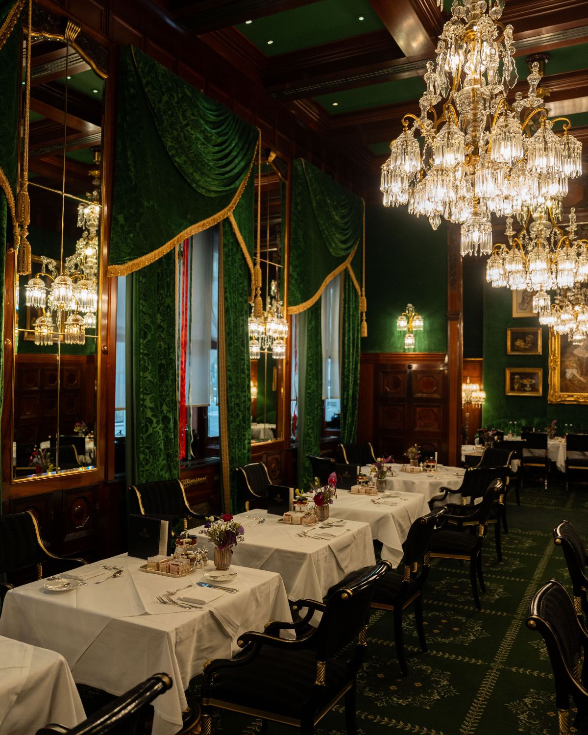 A green themed restaurant setting, with classical wooden paneling, green curtains and carpet, dark furniture, large crystal chandeliers and white tablecloths.