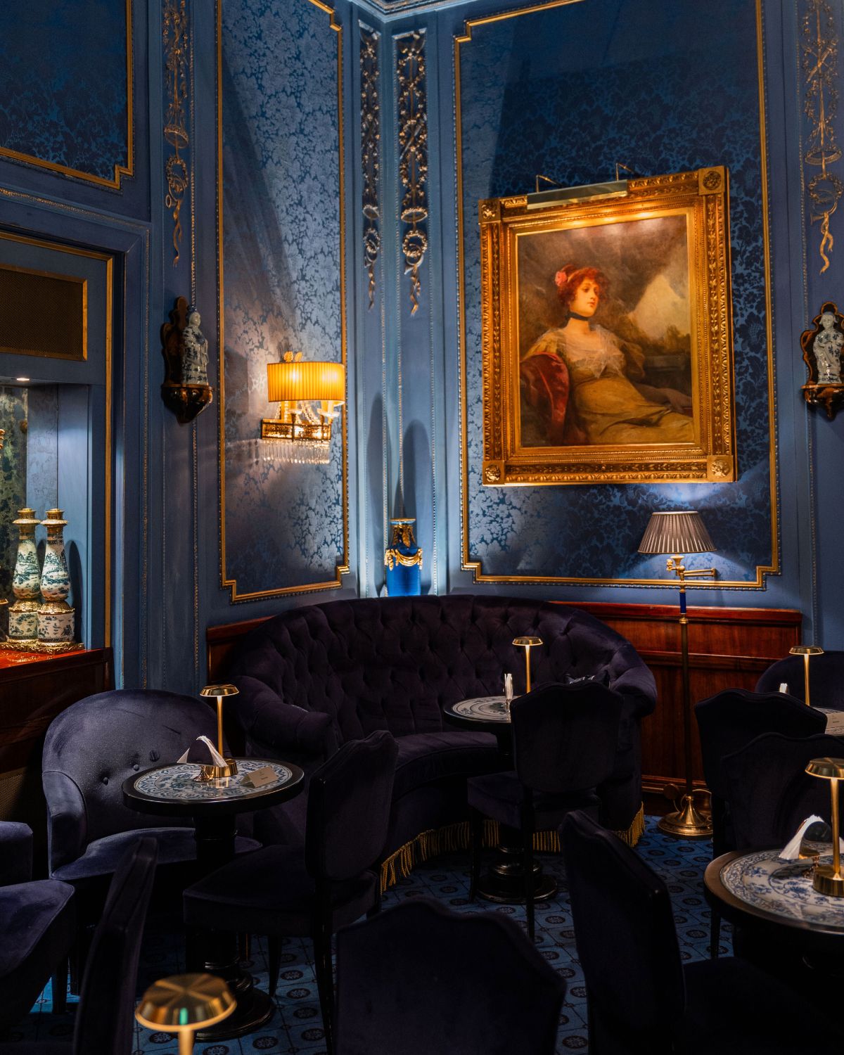 A blue themes hotel linge room, with gold paneling on the walls, a golden antique frame, dark blue velvet furniture and golden lamps.