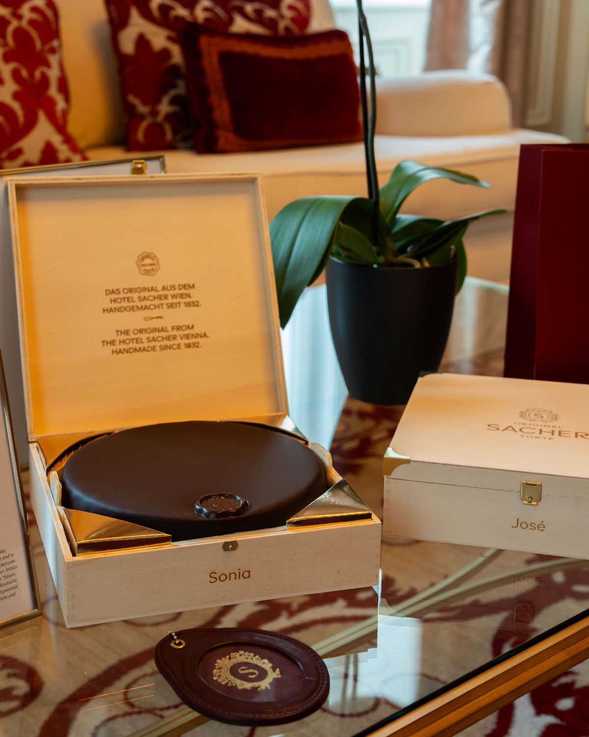 A chocolate cake inside a wooden box, on a glass center table, in front of a white sofa with red pillows.