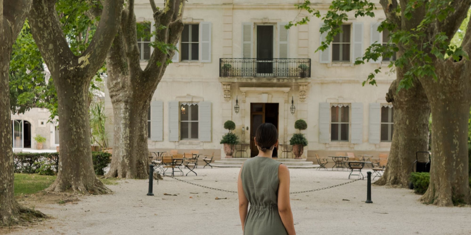 Cream house façade, with light blue shutters, outdoor wooden and ironed furniture, lack outdoor lamps and two vases with tailored bushes. Tall trees alongside a stone path, where a lady in a sage green dress stands.