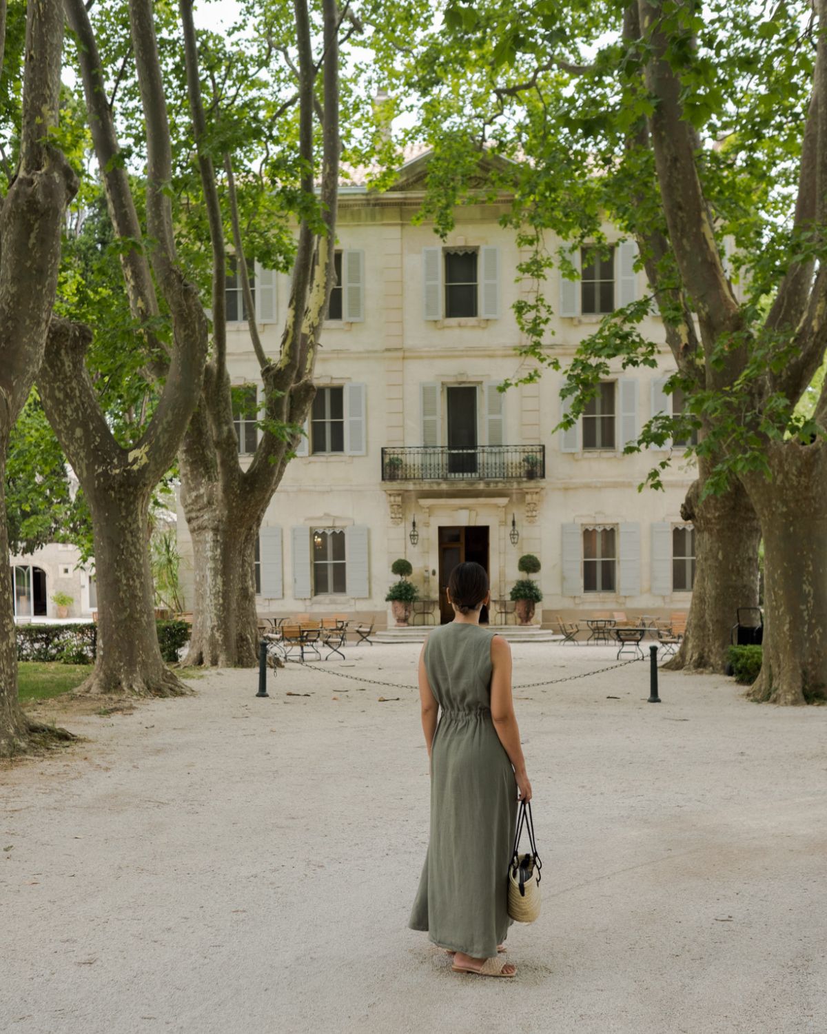 Cream house façade, with light blue shutters, outdoor wooden and ironed furniture, lack outdoor lamps and two vases with tailored bushes. Tall trees alongside a stone path, where a lady in a sage green dress stands.