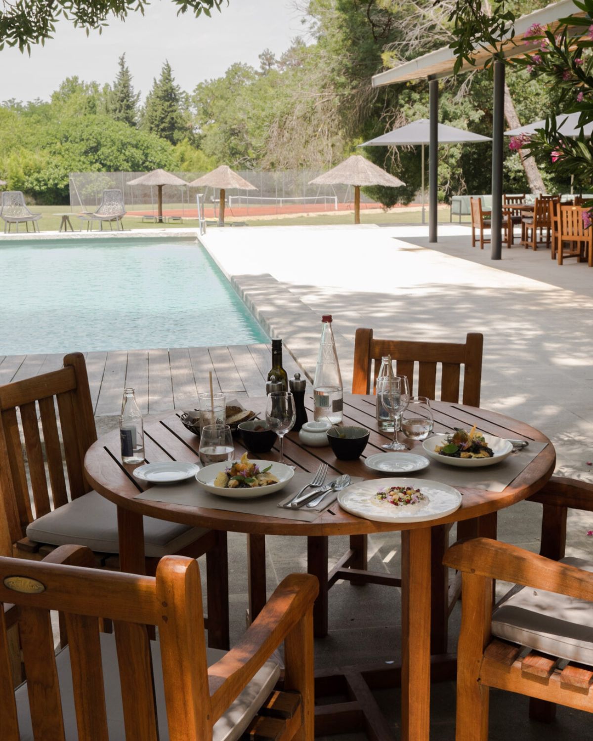 Light wooden table and chairs, over an exterior stone floor, facing a pool. White and black dishes on top of the table with diverse food, glasses and bottles.