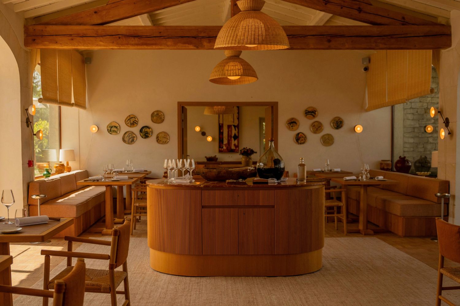 Warm toned indoor restaurant setting, with wooden beams and furniture. A center island with glasses and bottles. Decorative plates on the walls, hanging rattan lamps.