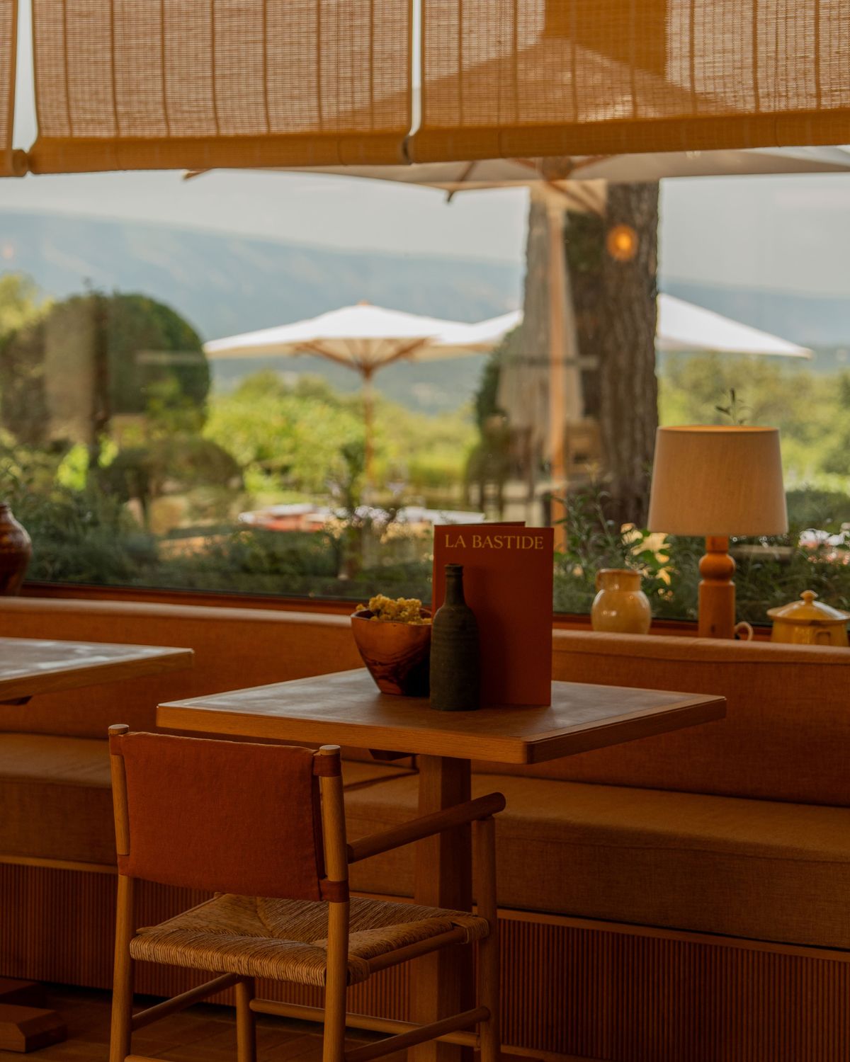 Warm indoor restaurant setting, with re and orange tables, blinds and chairs, overlooking white sunshades and green valleys.
