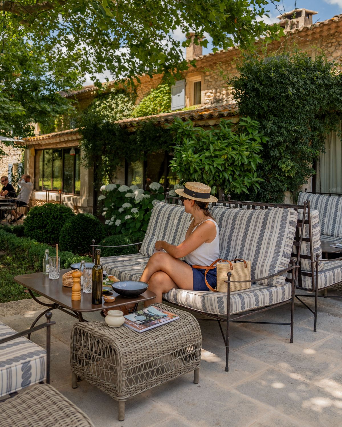 Cobble stoned house, with large windows and a stone pathway outside, under trees and bushes. Iron sofas and center tables, with white and blue cushions. A lady with a had standing in front of a restaurant table.