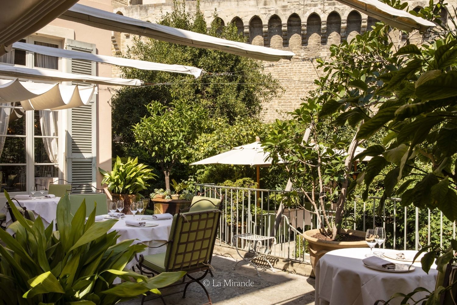 Outdoor restaurant setting, with pastel shutters, green iron chairs and white tablecloths. Green trees and bushes in the background.