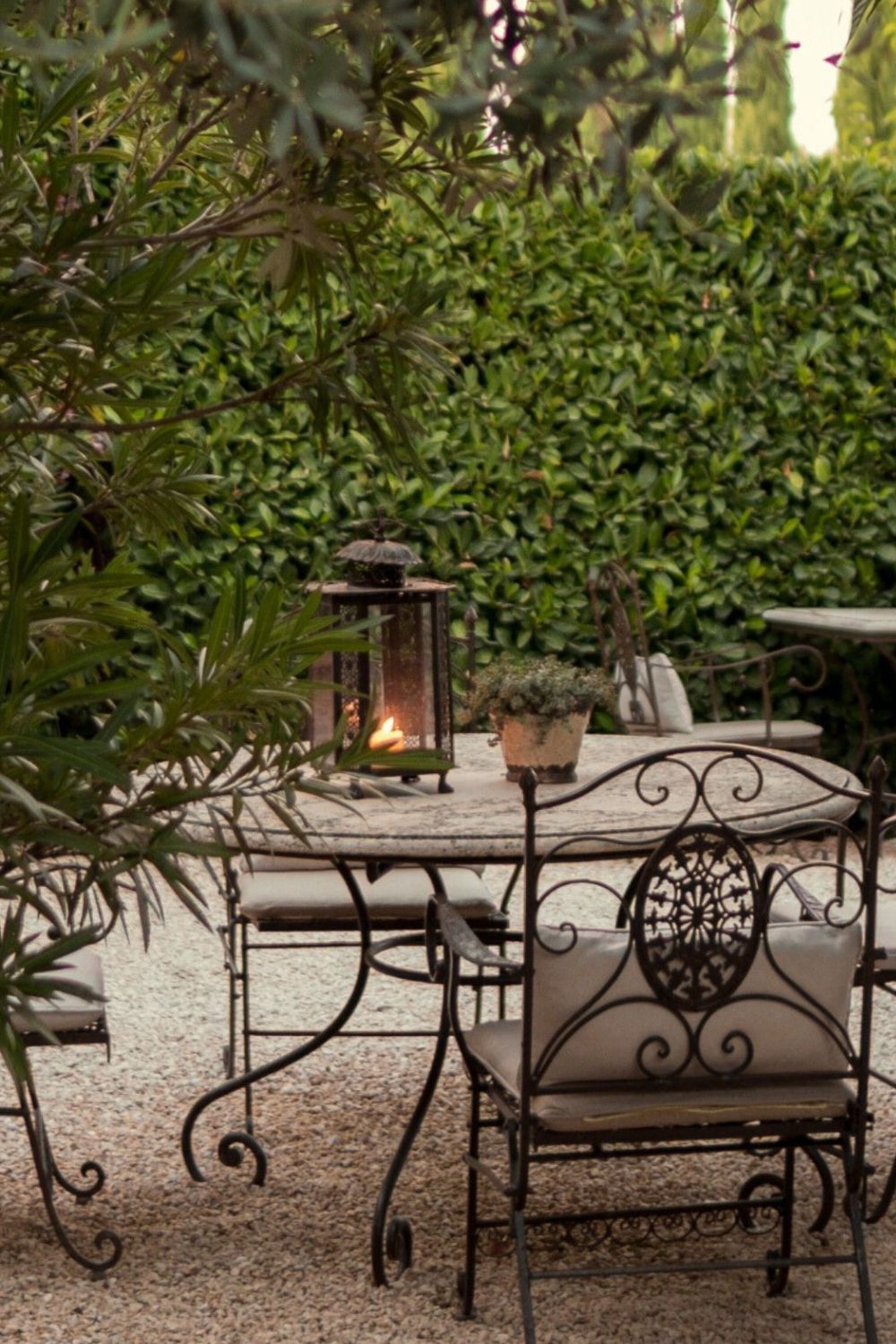 Dark iron garden furniture, with light cushions, a lit lantern on top of the table and a garden in the background.
