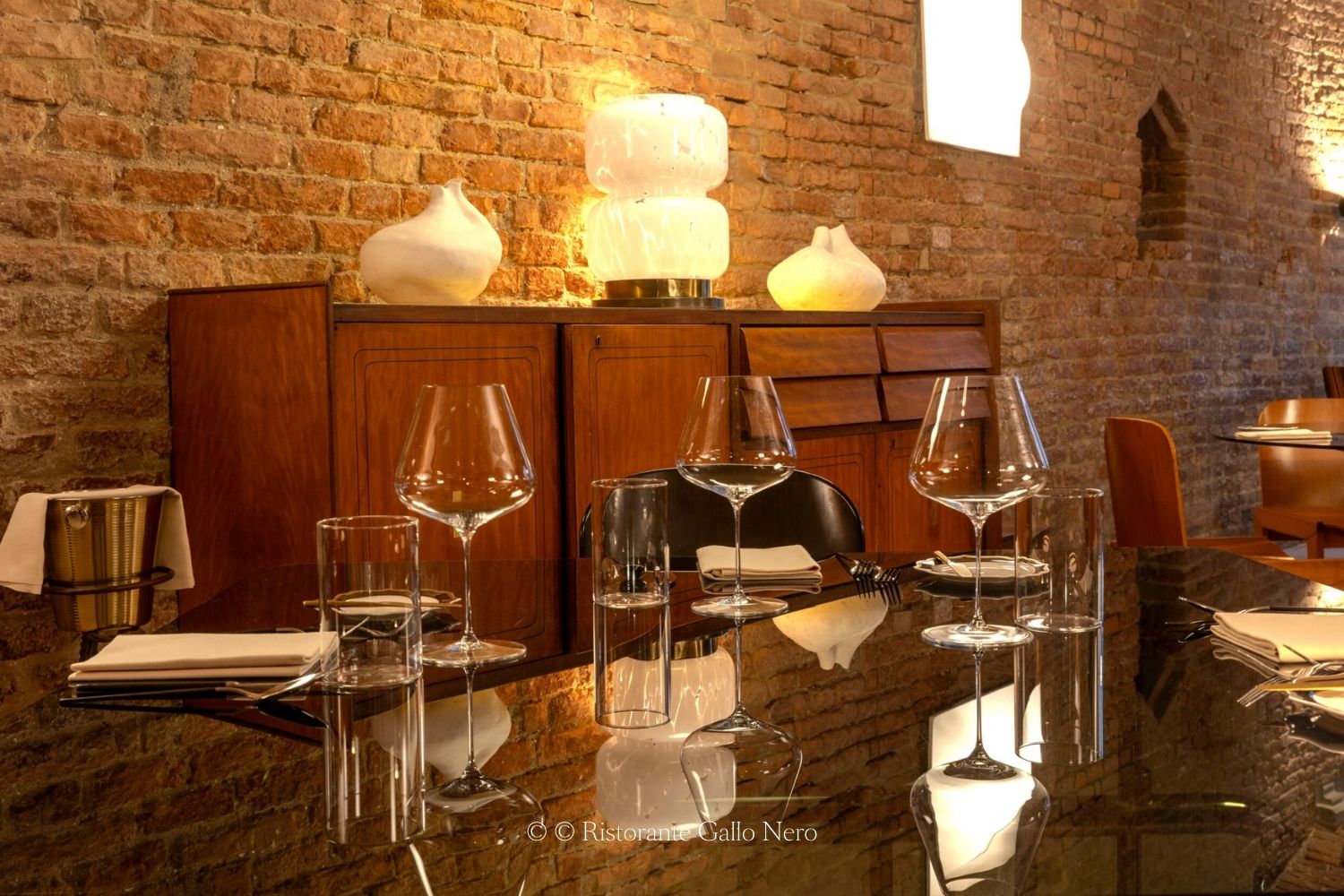 Exposed brick walls, with a glass table under several glasses and white napkins, in front of a vintage wooden cabinet.