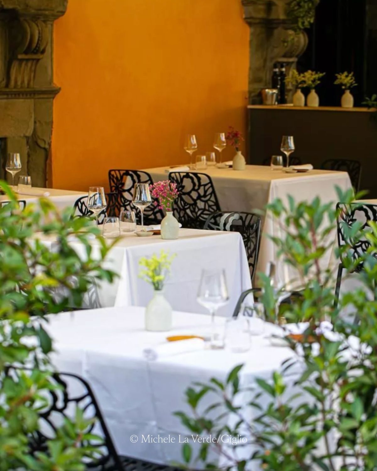 White tablecloths over several tables with black ornamented chairs, next to  bright orange wall and a stone fireplace.