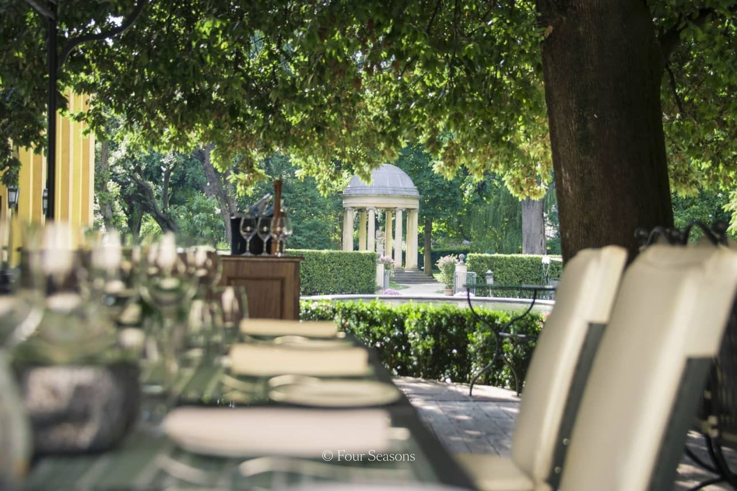 Outdoor restaurant setting with a large black table, with white linens and chairs, next to a wide oak tree and a garden in the back.