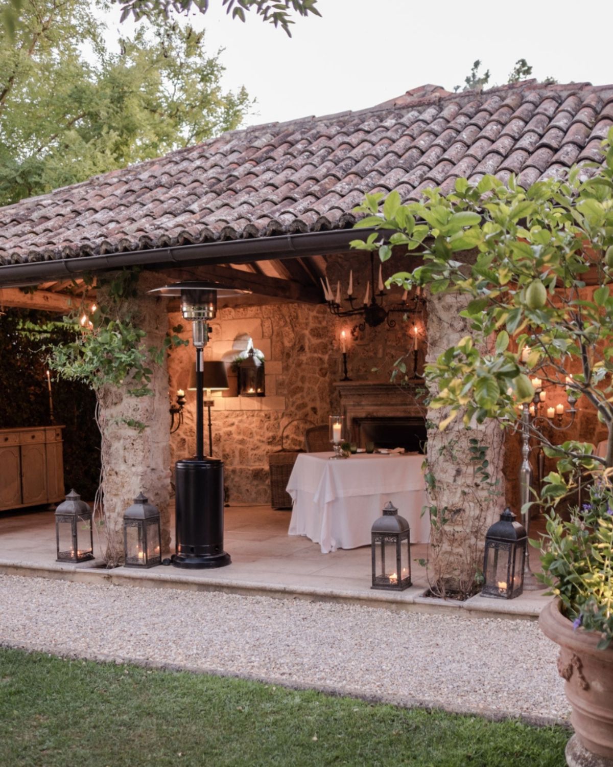 Stone open house with traditional roof, wooden beams, iron lamps and wisteria on the columns. A table with a white cloth facing a fireplace.