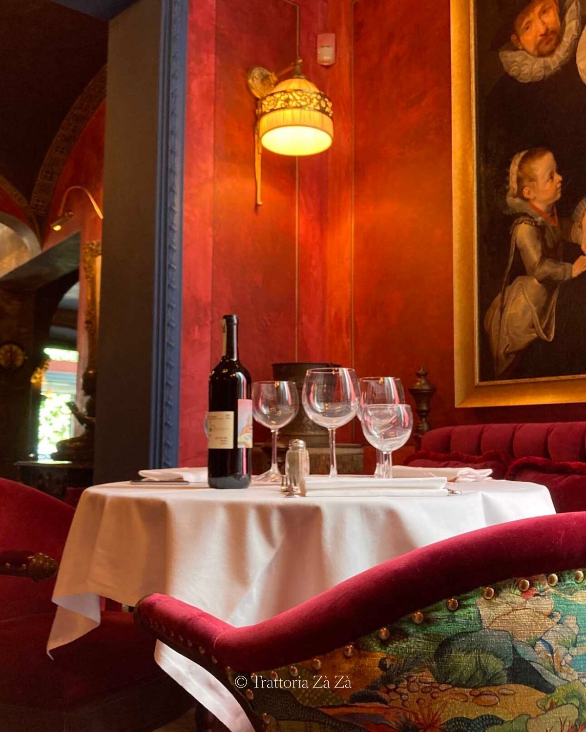 Restaurant interior setting, with bright red walls and sofas, white linens, glasses and a bottle of wine. Blue rims over the doors and an old golden painting.
