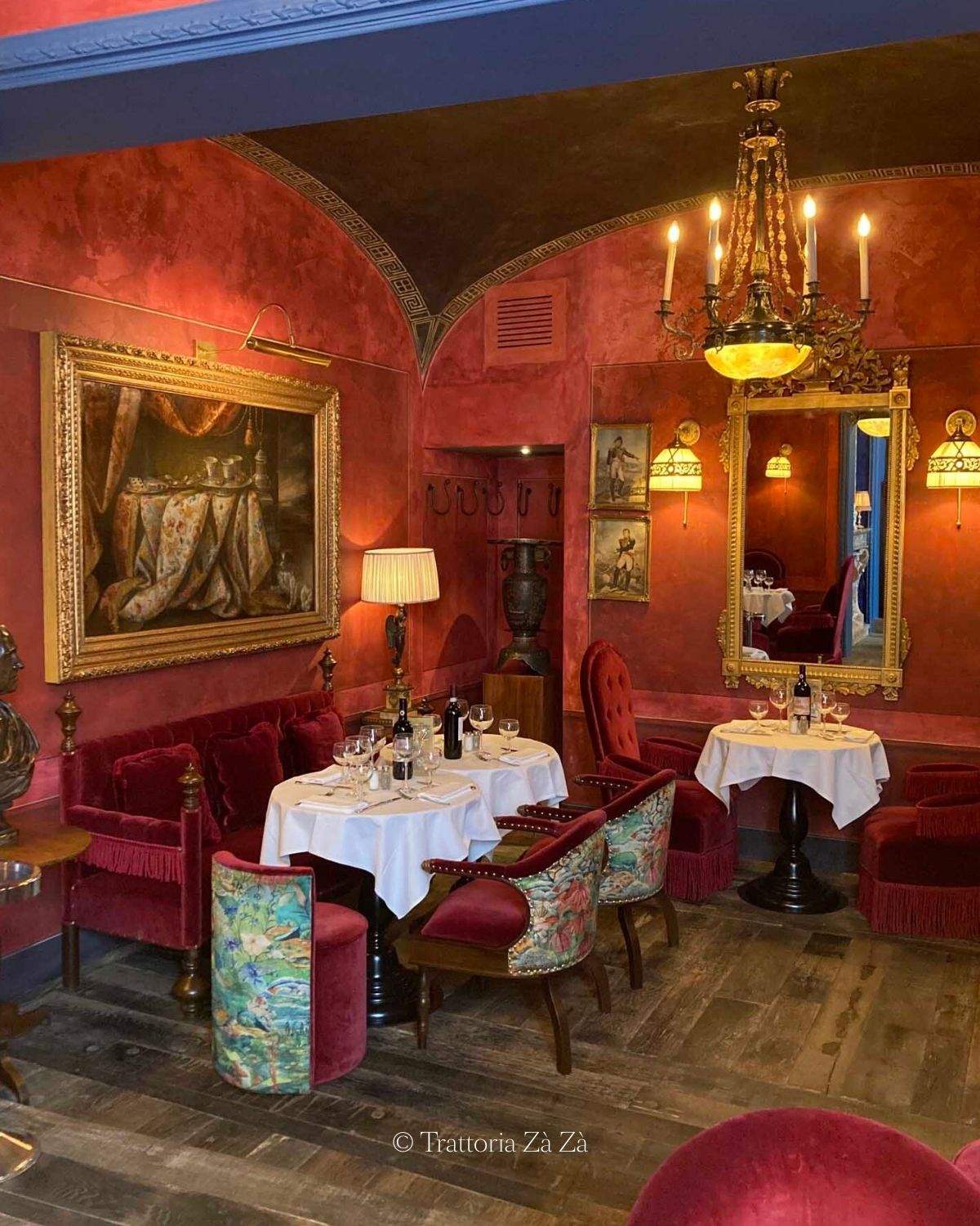 Restaurant interior setting, with bright red walls and sofas, white linens, glasses and a bottle of wine. Blue rims over the doors and old golden paintings and mirrors.